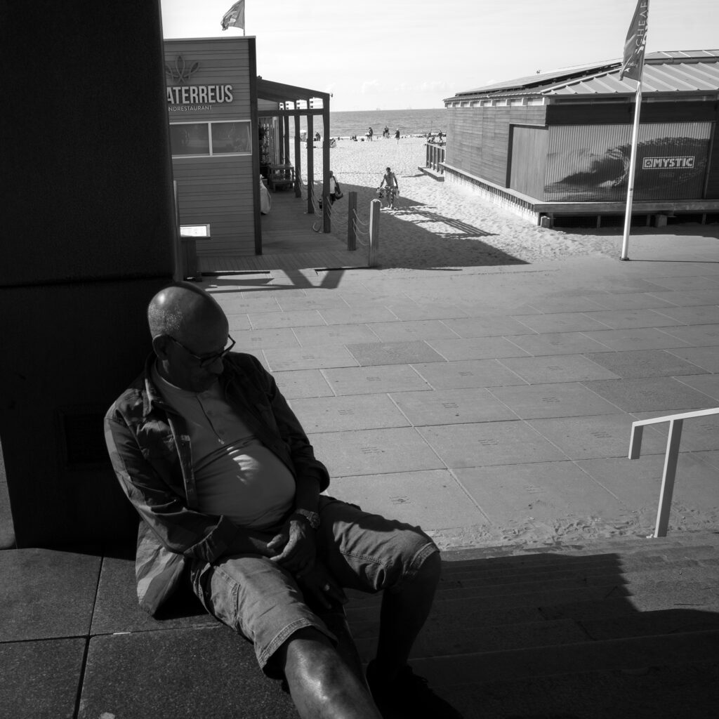 Person resting in the shade on Scheveningen&rsquo;s Noorderstrand promenade near De Waterreus, with the beach and North Sea beyond.