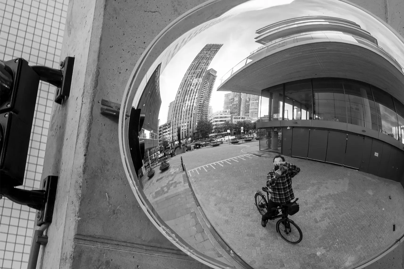 Point ’n Shoot | A black-and-white convex mirror reflection showing Loyd ’t Hart on his bicycle, camera raised, with modern Rotterdam buildings and an open plaza in the background.