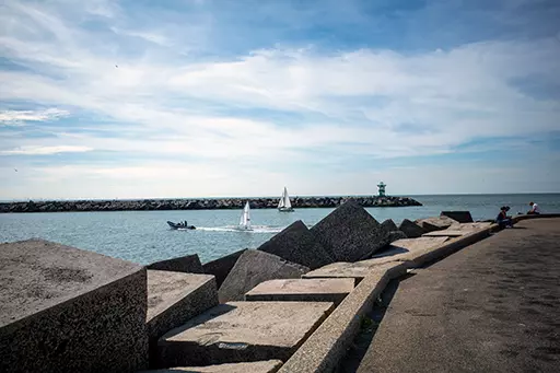 Sailboats entering the harbor near the northern harbor head of Scheveningen with concrete breakwater blocks in the foreground.