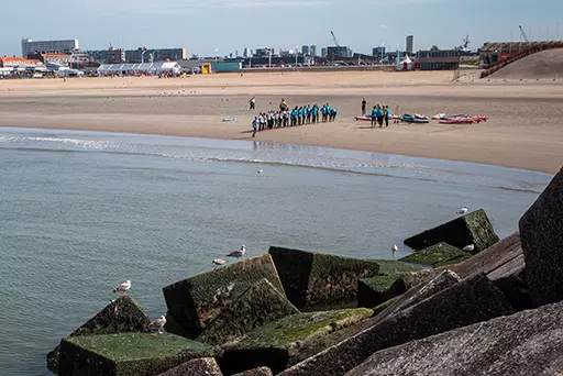 View from the northern harbor head of Scheveningen overlooking the beach, breakwater blocks, and people gathered near the shoreline.